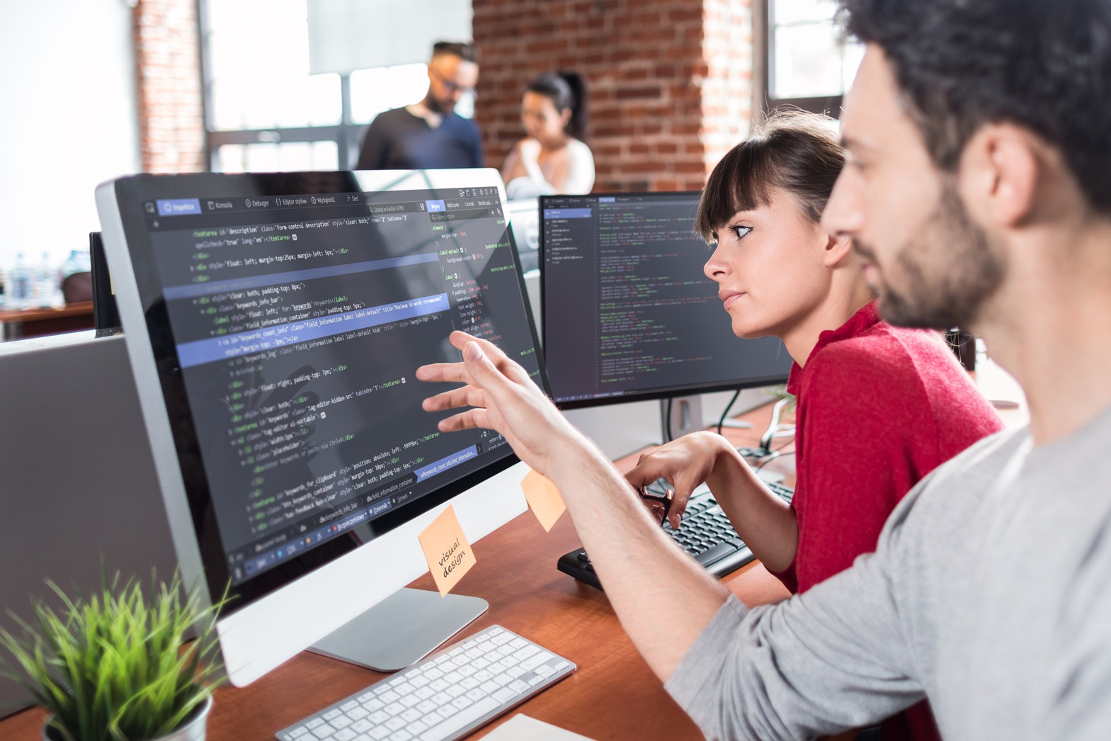 Developers collaborate on coding tasks at a modern workspace, with one pointing out elements on the screen while the other observes.