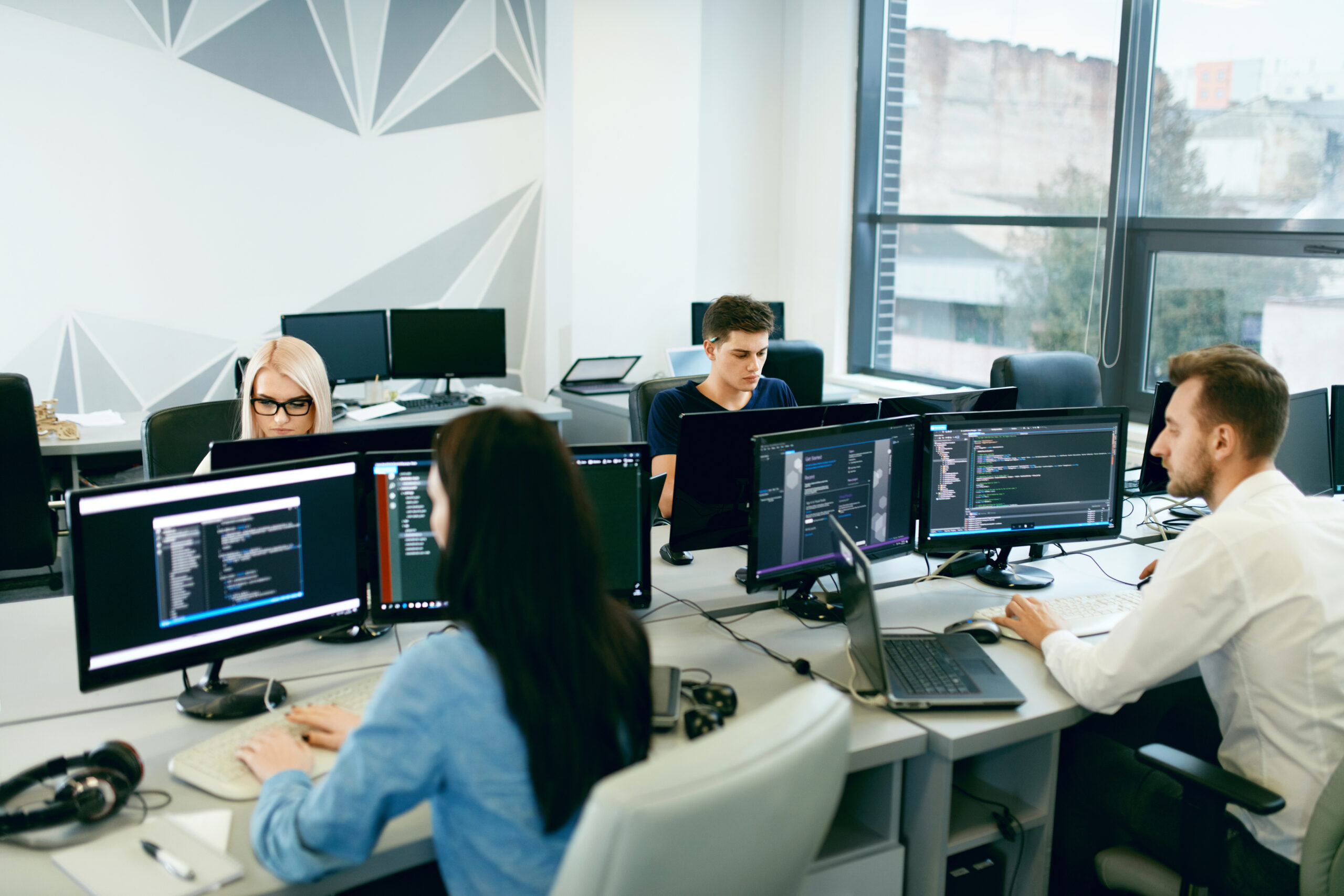 Four professionals are focused on coding at a modern office workspace, featuring multiple monitors and a bright, open environment.