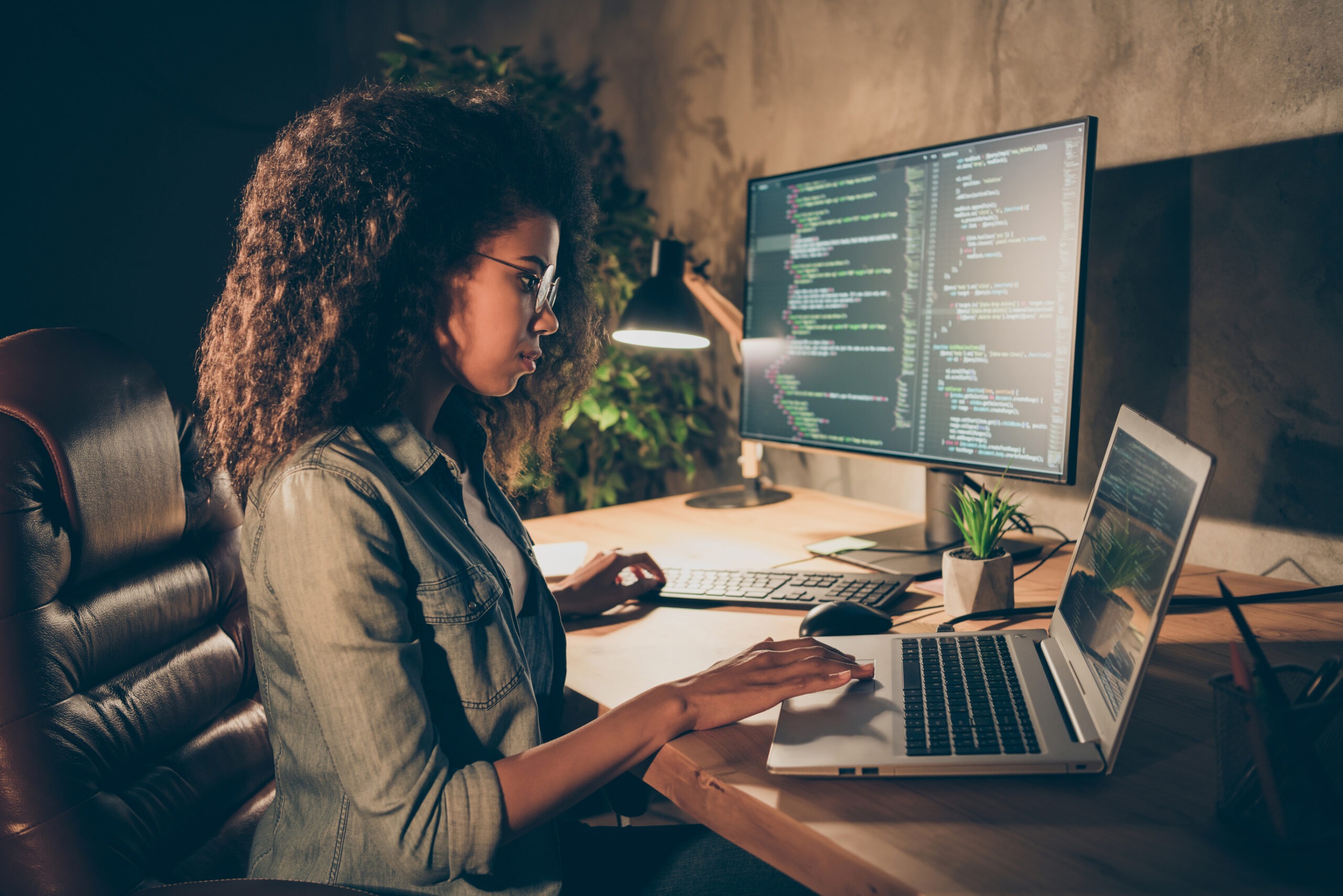A woman with curly hair works intently on a laptop in a dimly lit room, surrounded by coding on a large monitor and a small plant.