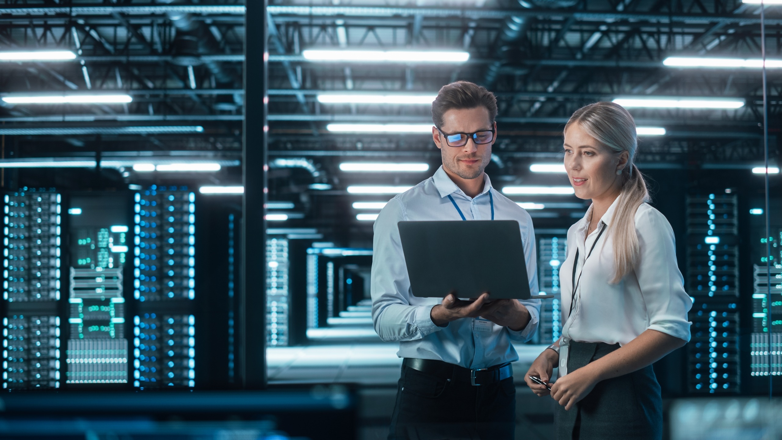 Technicians discuss data management while reviewing information on a laptop in a modern server room, emphasizing teamwork and technology.