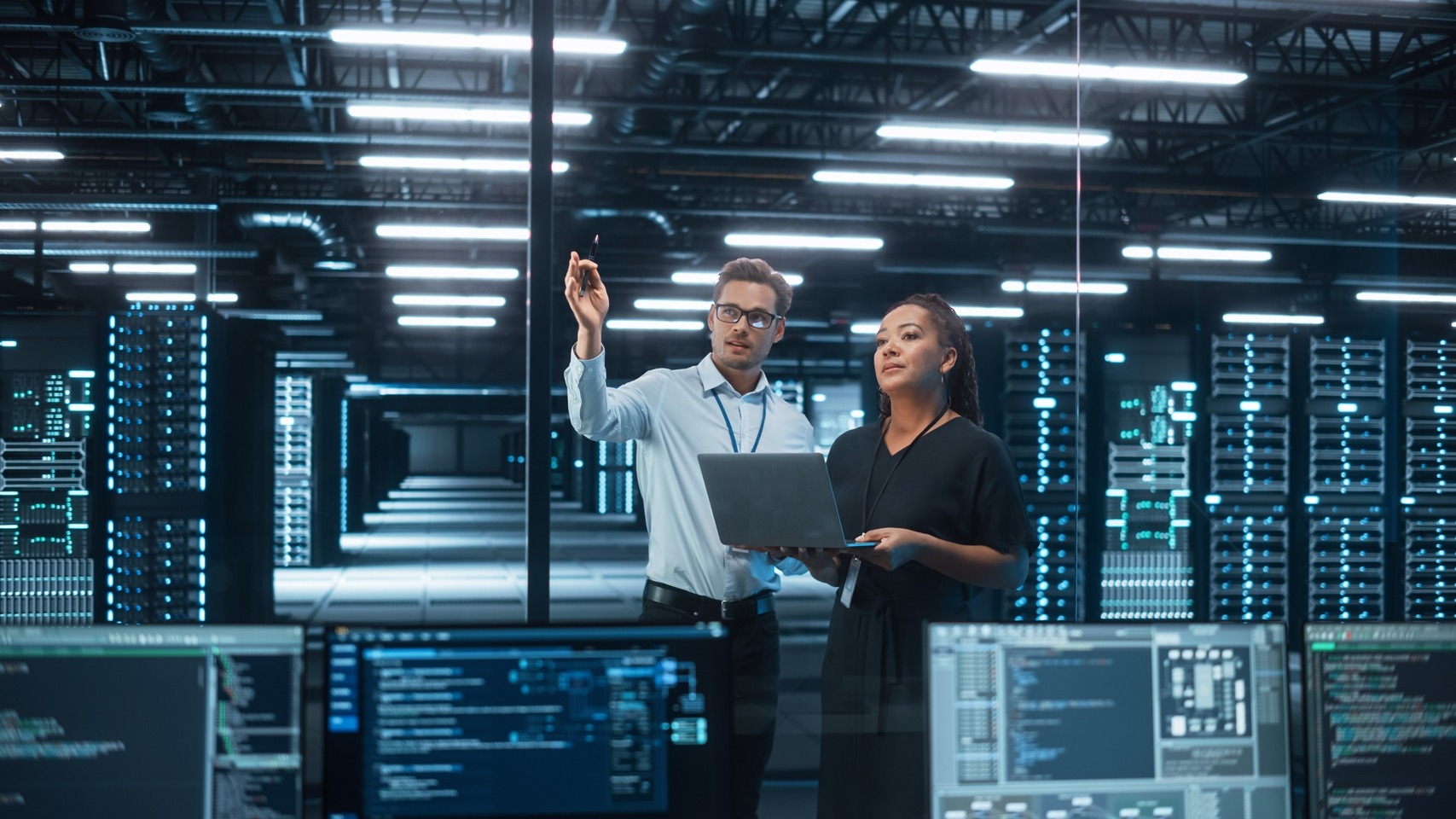 Two professionals discuss a project in a modern data center, with rows of servers illuminated behind them, showcasing an advanced tech environment.