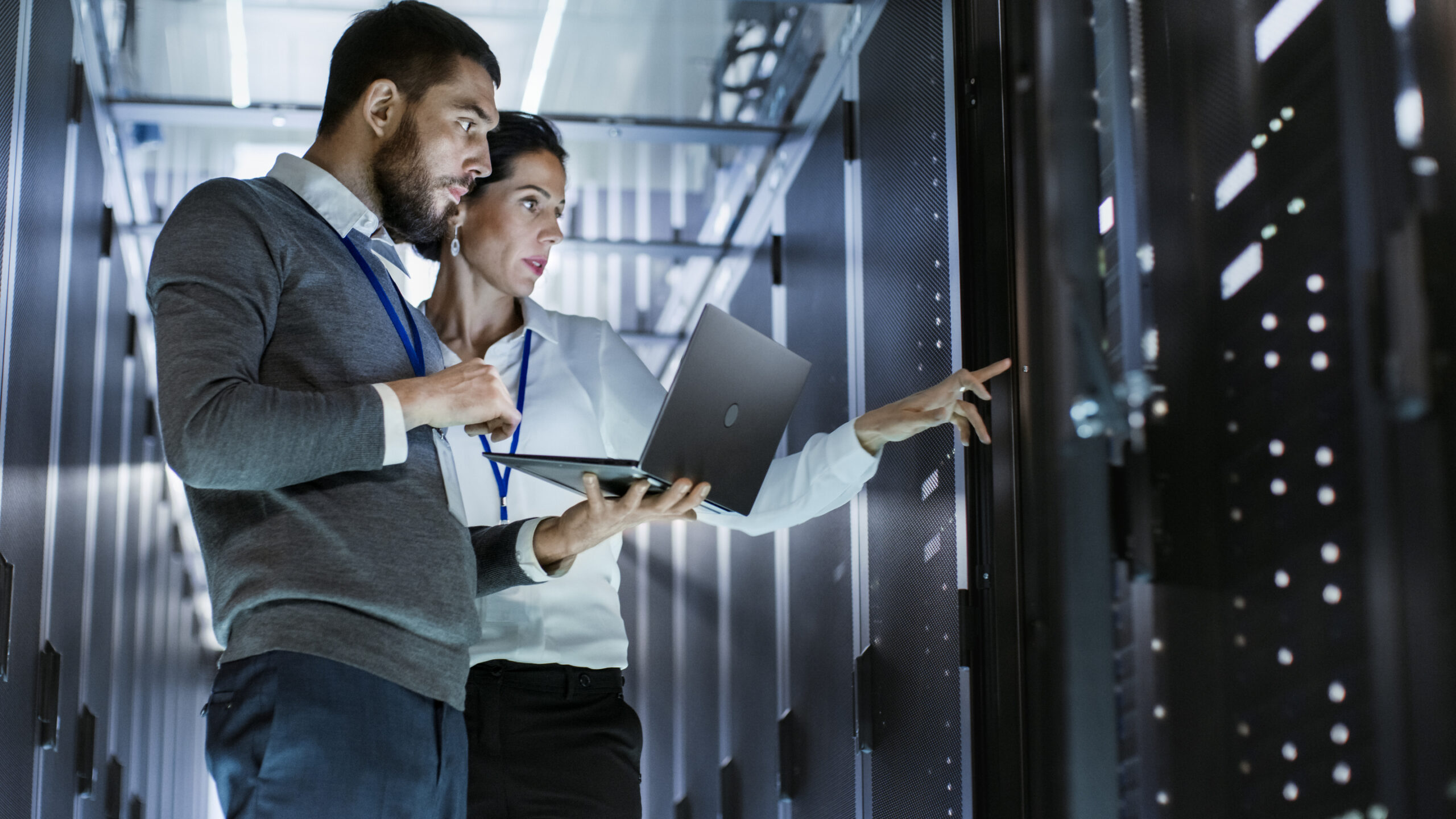 Professionals in a data center, analyzing information on a laptop while discussing server configurations, highlighting collaboration in tech environments.