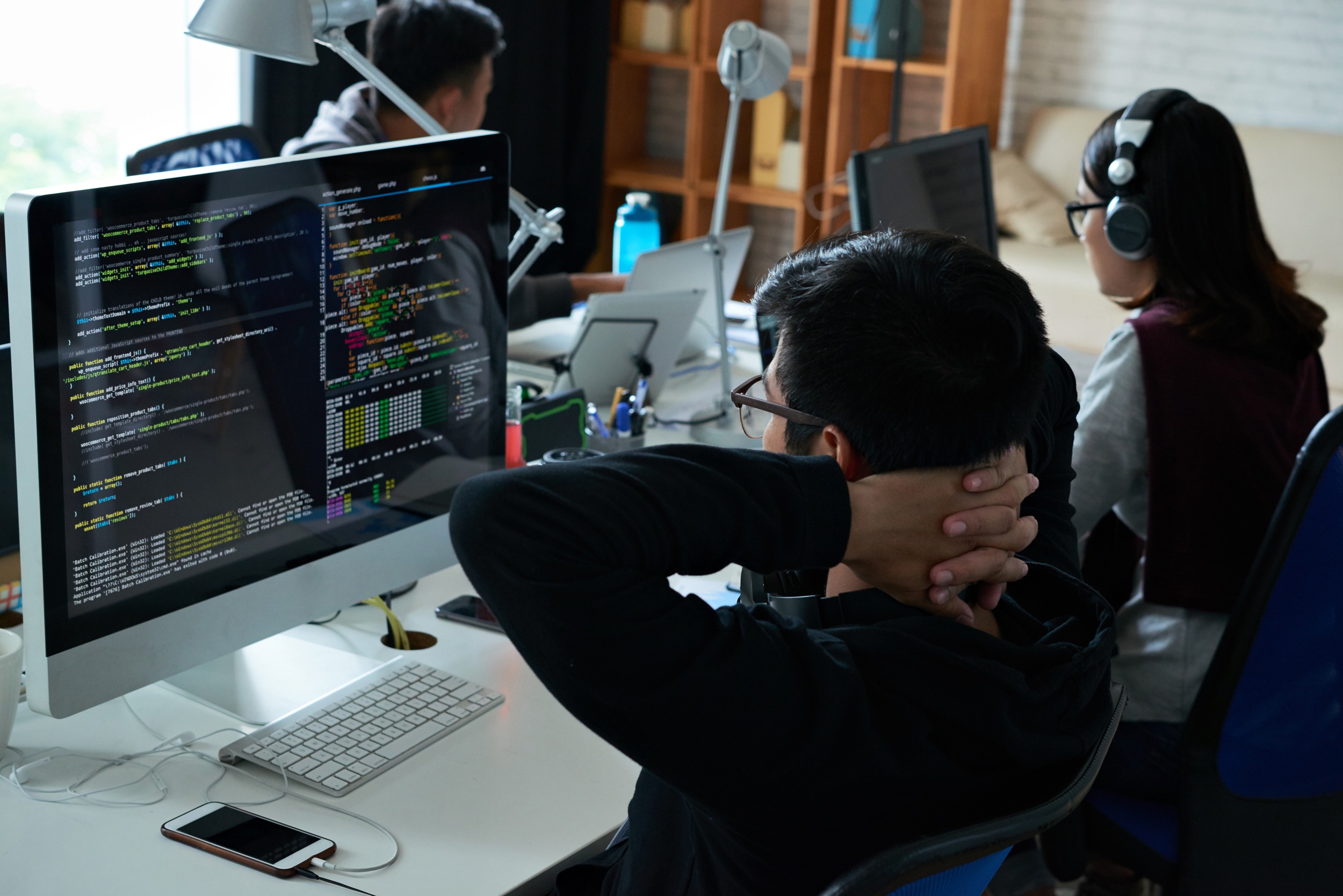 A programmer sits at a desk with hands behind their head, analyzing code on a large monitor. Colleagues work on laptops in a modern office environment.