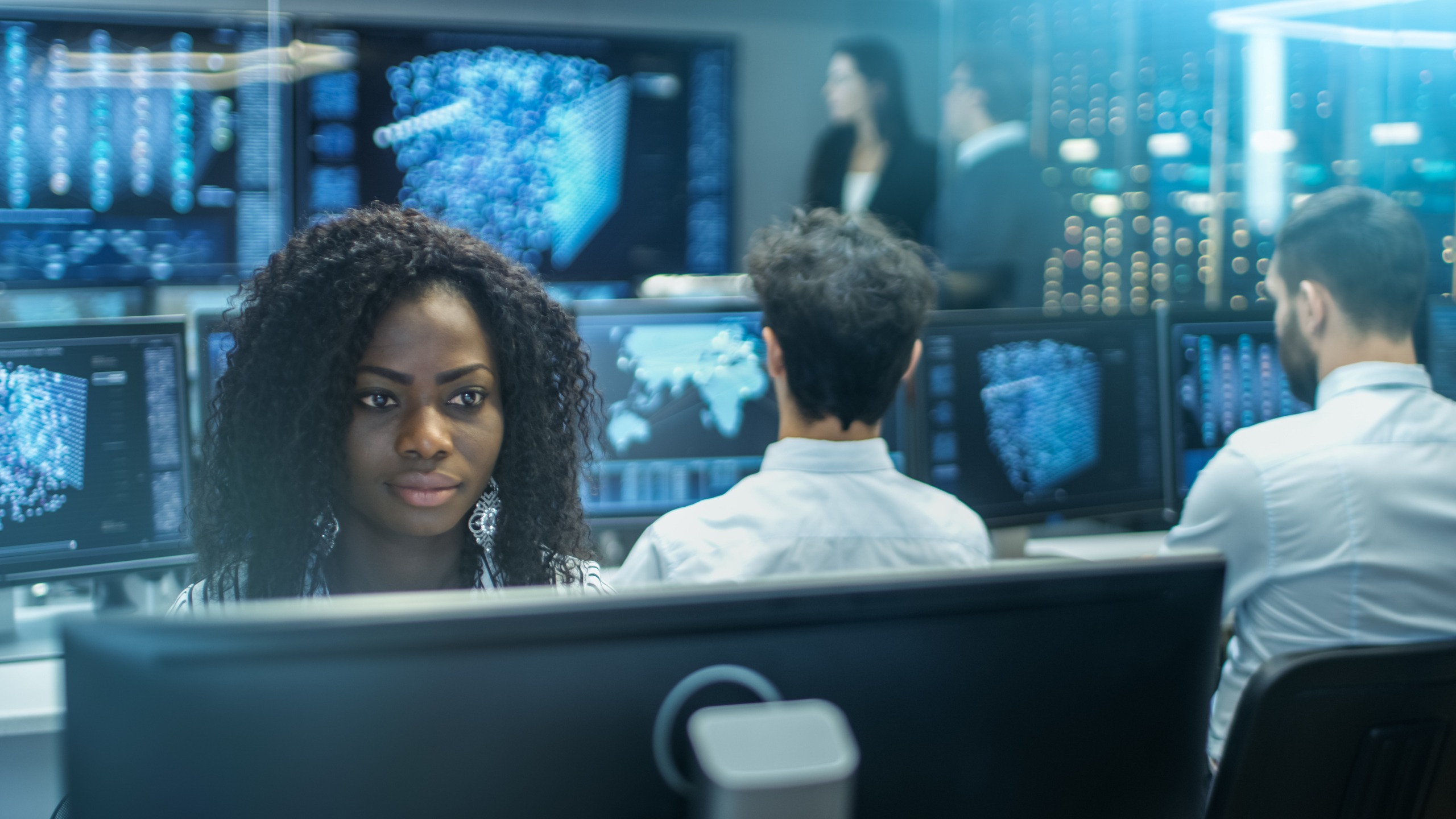 A focused data analyst, her curly hair framing her face, works intently at a computer in a high-tech office, surrounded by colleagues and digital displays.